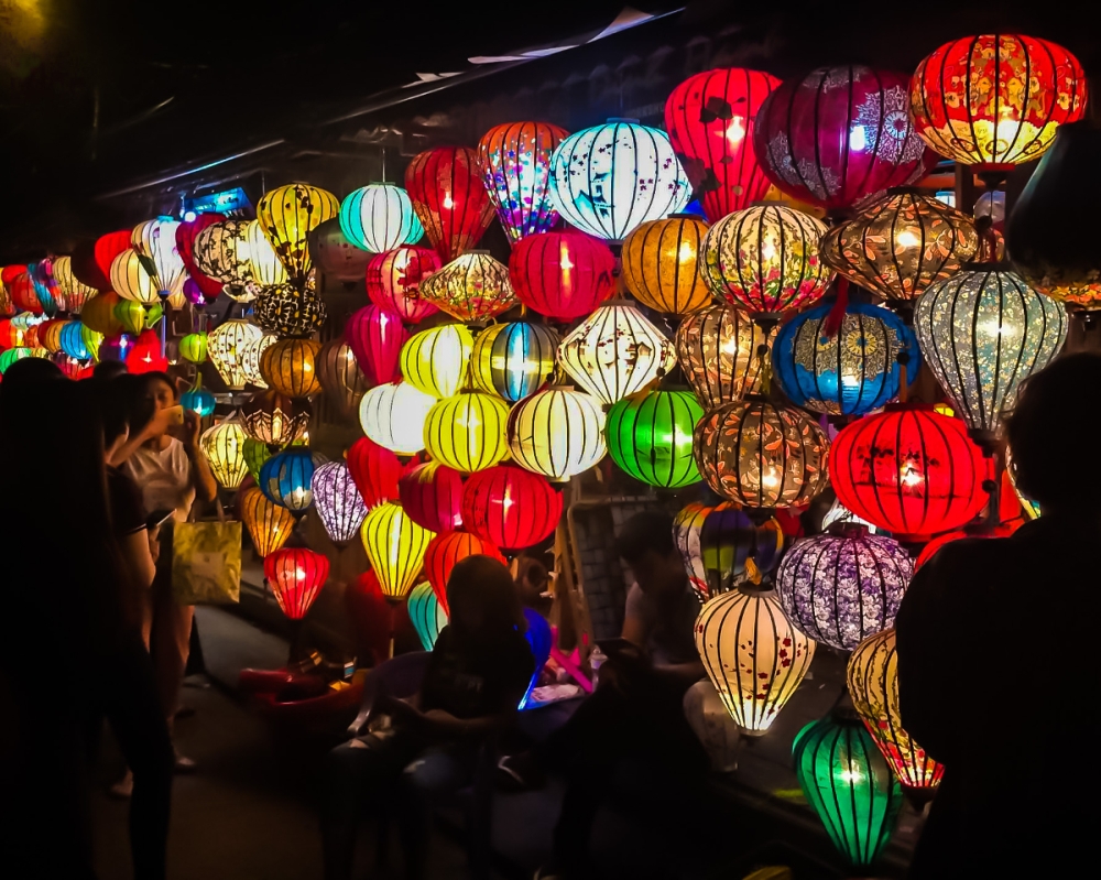 A night photo of a wall of bright paper lanterns in multiple colors, all of them with a light burning inside. A person is dimly visible sitting in front of them, a silhouette with the bright lanterns behind them.