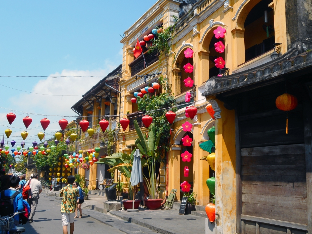 A view along a row of buildings in Hoi An, all one or two stories tall, with a flat-topped facade. They're painted in shades of light orange and are very colorful with chains of mostly red paper lanterns hanging down their facades and draped across and above the street as well.
