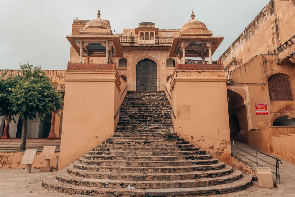 Grand entrance to a building in pinkish stone. A central stairway up, wider at the bottom than further up. A small tower on either side of the stairway, each with a small domed roof above its top - guard towers, maybe? Not much of the building at the top of the stairway is visible, but it has a large arched doorway.