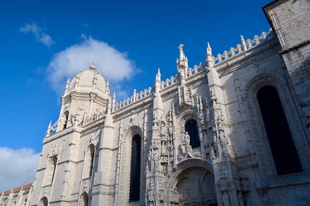 A partial view of the Jeronimos Monastery shows a very ornate arched entrance, with lots of decorative embellishment around and above it. Along the roofline are filigreed crenellations and a corner of the building has a dome-topped little tower.