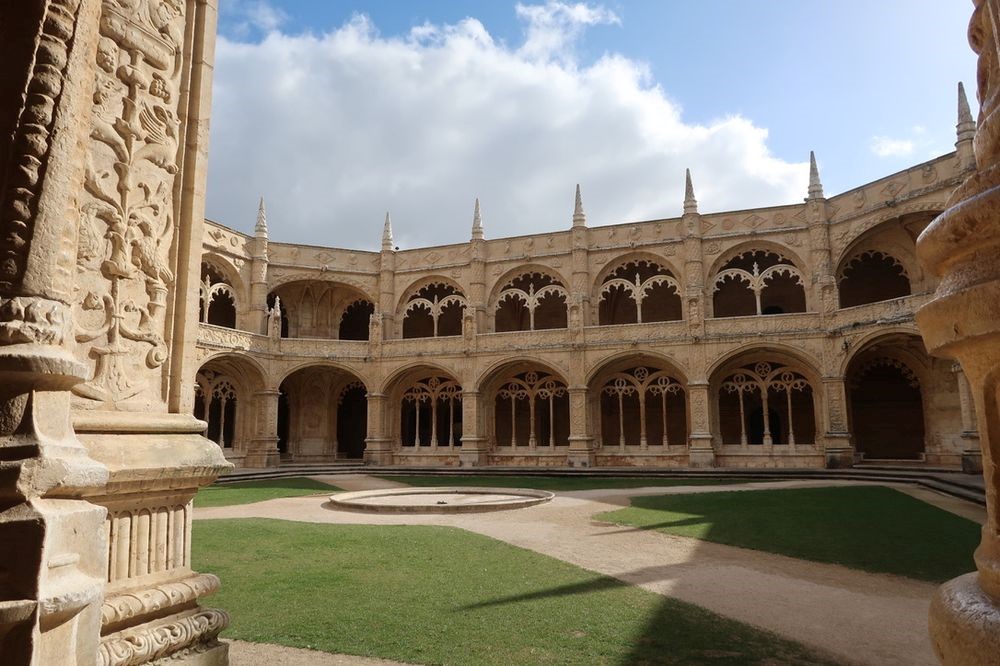 A couryard that looks round or multi-sided, with flat green grass on the ground. The walls are 2 stories and lined with arches held up by square and highly-carved pillars. Inside each arch are two windows set further back, each of which has a decorative arch over it.