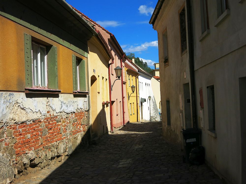 A cobbled street curves ahead, lined by simple one and two-story houses, each covered in plaster and painted a pastel shade. The nearest house on the left is quite damaged: the plaster on the bottom half of the wall has come off and the brick underneath is visible.
