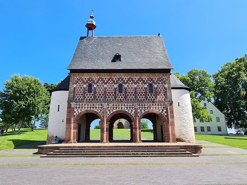 A small building standing apart from other buildings, the gatehouse at Lorsch has two stories. The ground floor is open, with 3 arches front and back, so, from this front view, you can see through the building. The upper story has 3 small windows. The facade has rather intricate stonework, with a checkerboard pattern of red and light brown bricks. A half-round structure has been added at each end.