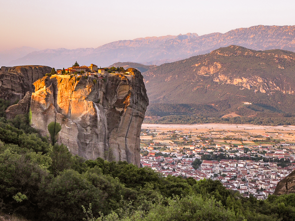 Left side of the photo an outcropping of rock. Right side: a view down a valley to a city below, with mountains behind. Buildings on the flat top of the outcropping - whitish with red roofs - look extremely small in relation to the size of the cliff below them.