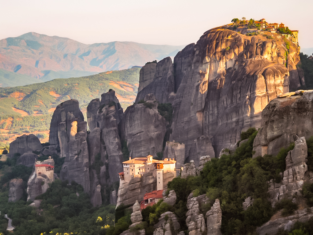 High rocks like columns. Front center, low in the photo, is a monastery, in white plaster with a red roof, on a  small outcropping. Behind it is a far taller rock, and at the very top right of the photo, more buildings are visible, looking tiny on the top of the rock. Mountains in the distance behind.