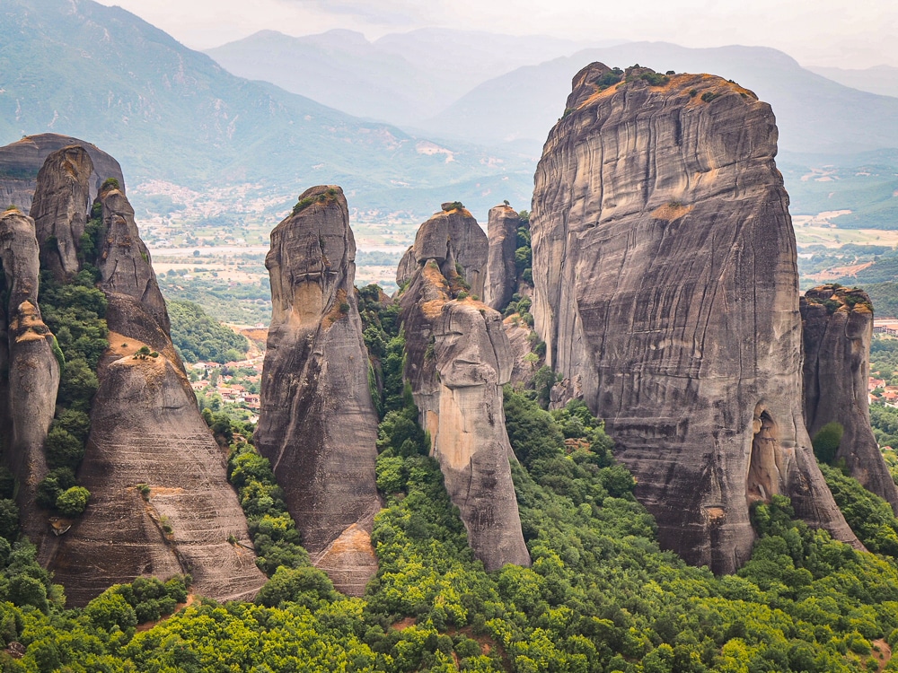 5 vertical columns of rock in a row, with forest growing between them. Too far to see whether any monasteries on on any of them. Beyond and between them mountains are visible.