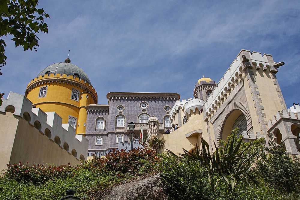 Looking up at the palace. At the left-hand end a round tower with a round dome on top is painted mustard yellow. The middle part of the building is flat-roofed and painted grey, with arched windows. A piece of a wall projects down the hill toward the camera at the right, complete with crenellations.