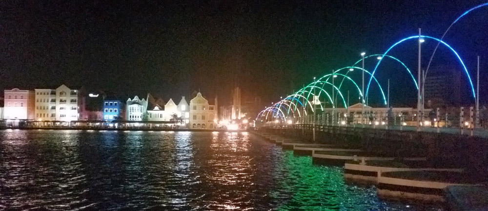 A night-time view over the river. In the distance, a row of colorful buildings on the riverbank, all lit from below. On the right, the pontoon bridge has arches of neon lights over it in a range of colors.