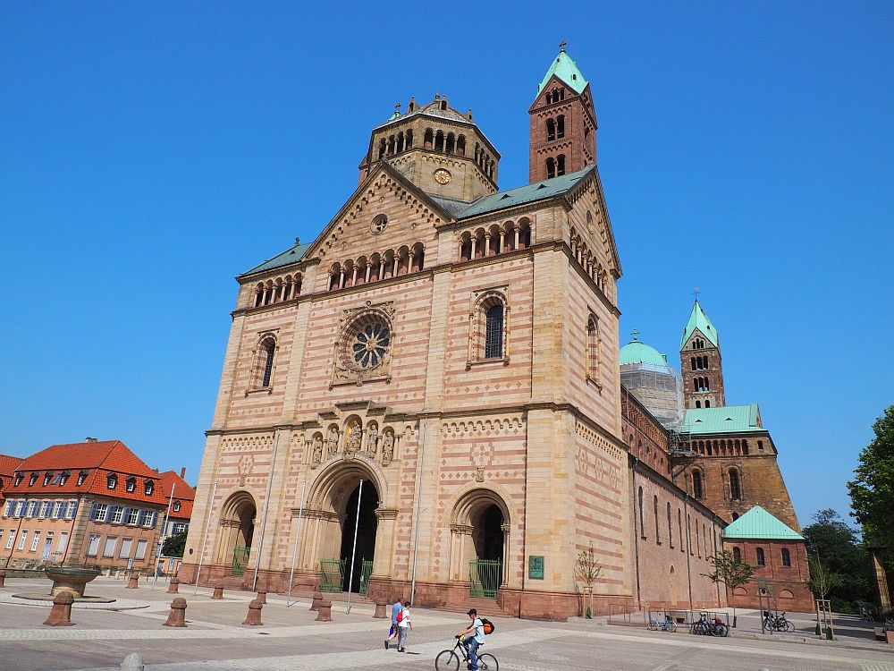 A view of Speyer Cathedral from an angle. The front is flat and relatively unadorned in stripes of red brick and lighter brick. An 8-sided cupola on top of it and 2 towers are visible, one on the near side of the cupola and one at the back of the church. 