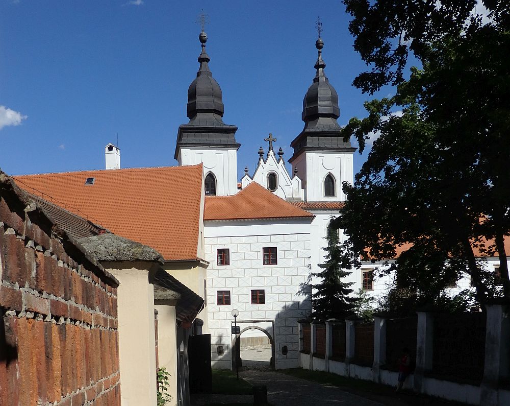 A narrow street with walls along the sides leads to a white building with an archway in it. Behind that, part of the Basilica is visible: 2 white towers with dark grey roofs that have elongated domes. One has an orb at the top, the other a cross. The roofline between the towers is also visible, and has 5 small peaks, each with a cross on it.