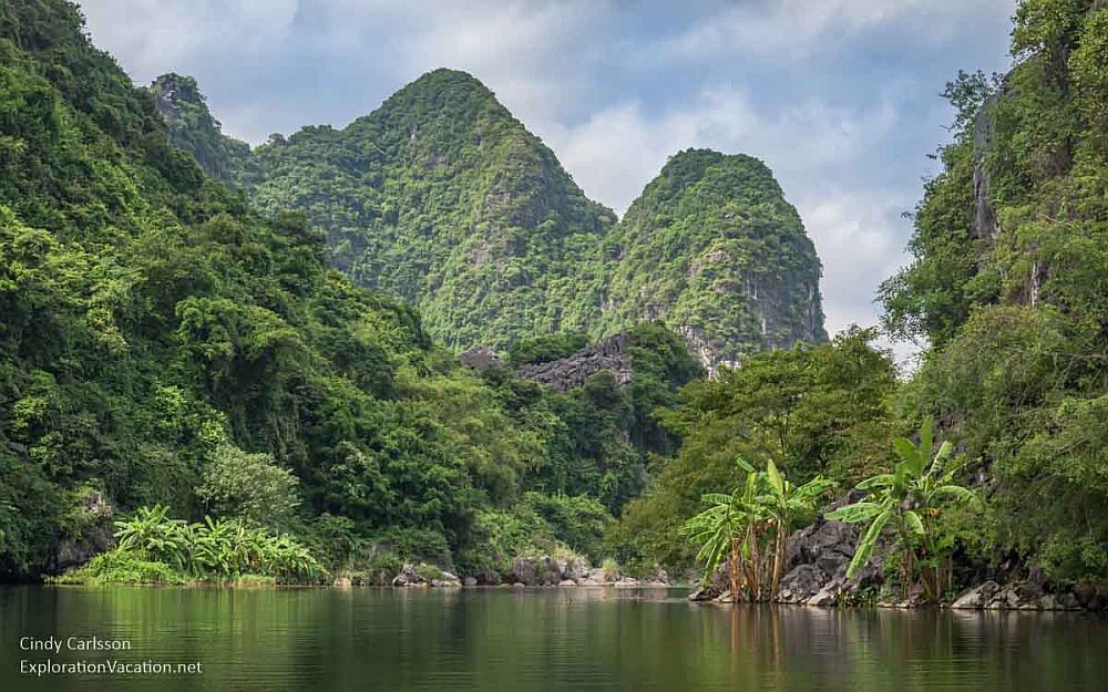 From a river, looking onto the land, which is covered with tropical greenery. Two very steep mountains rise behind the riverbank.