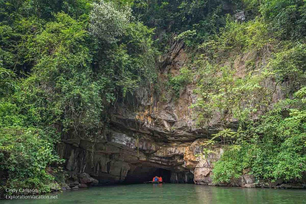 In a rocky cliff with sparse plants growing on it, the river enters a low tunnel. A boat can be seen entering the tunnel.