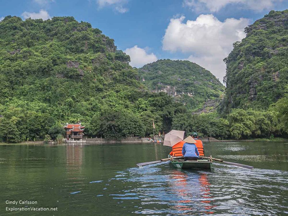 From the river, looking toward land. Three greenery-covered round hills and one small pagoda on the riverbank. Nearby, in the center of the picture, the back of a boat. A person in a pointed hat rows at the rear of the boat. Two people wearing orange life jackets sit in front of the rower, one of who carries an unbrella.