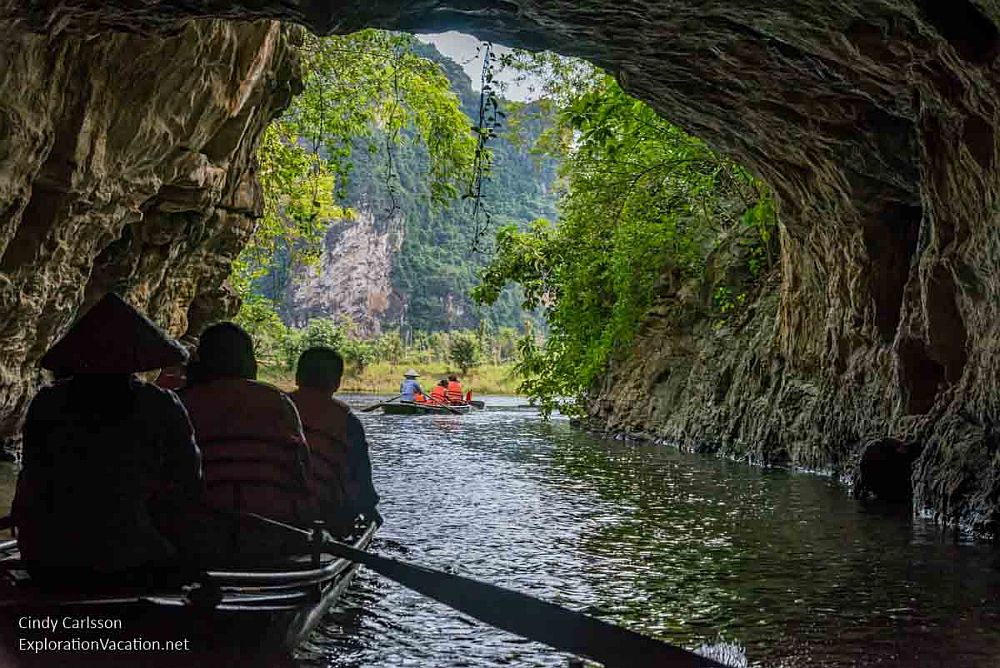 A photo taken inside one of the tunnels looking out toward the open river. In front is another boat seen from behind. The rower sits at the rear and wears a pointed hat. Two people sit in front of him/her. Outside the tunnel, in the light, another such boat is visible.