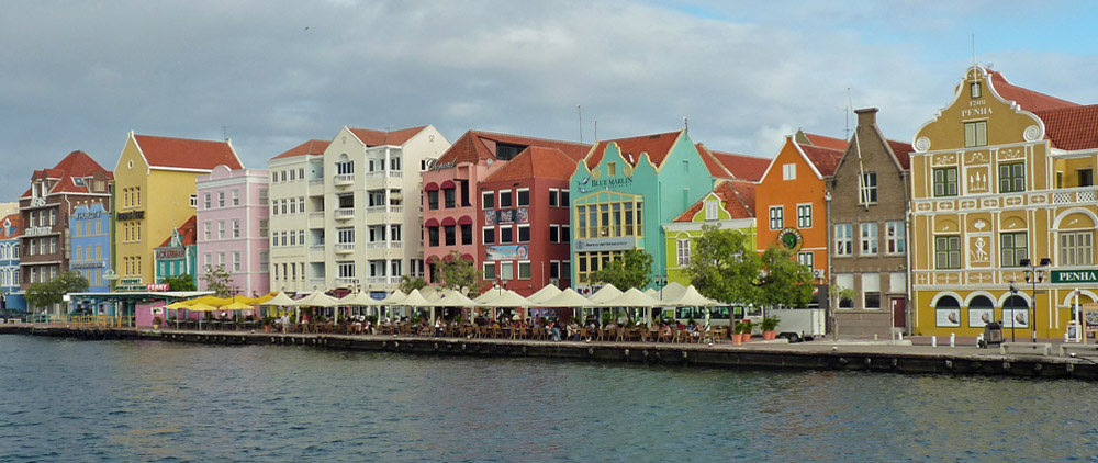 A row of houses on the other side of a river: very much a Dutch style of facade, tall and narrow with a variety of different roof shapes. Unlike in the Netherlands, though, they are painted in lots of different colors. In front of them, between the water and the buildings: outdoor cafe seating with umbrellas.