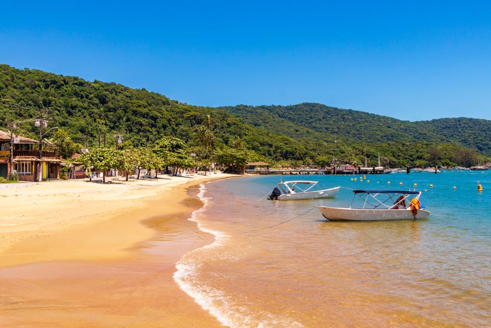 Looking along a long sandy beach. Some low buildings and trees along the left side, on the beach, and two boats moored in the shallow water. Hills covered in greenery behind the beach.