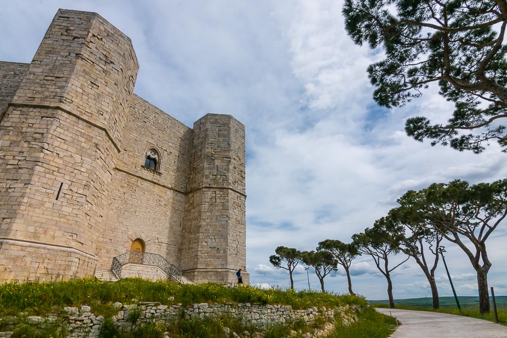 A view of one of the sides of Castel del Monte, showing two octagonal towers flanking one flat wall with a simple door at the base up a short stairway and a single window above the door on the upper floor.