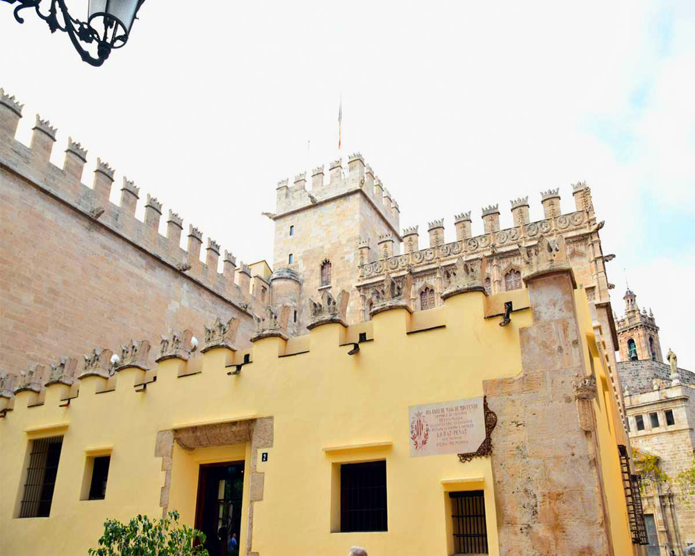 The photo looks up at the building, and shows the roof lines: it has crenellations, but more ornate than the usual castle crenellations: these have decorative carvings. the lower part of the building is painted pastel yellow.