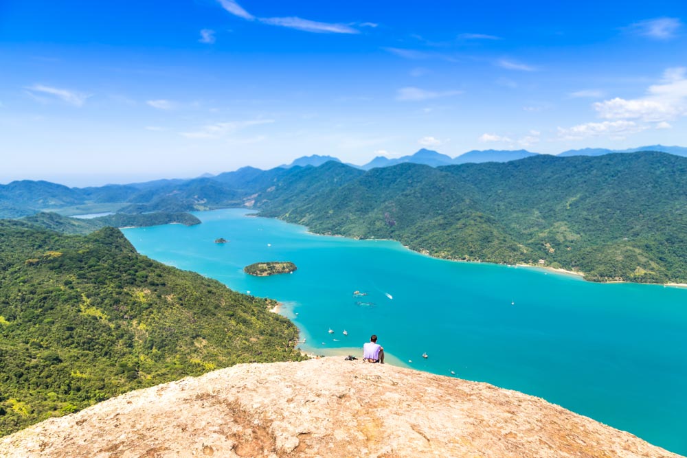 Mountains on either side of a long narrow body of very blue water. A person sits facing the water, back to the camera.