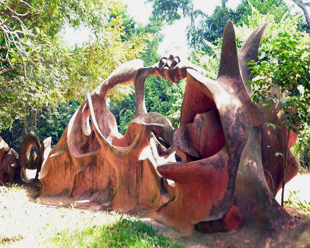 In a shaded grove, a large abstract artwork that has rather organic shapes with an arch extending over them.
