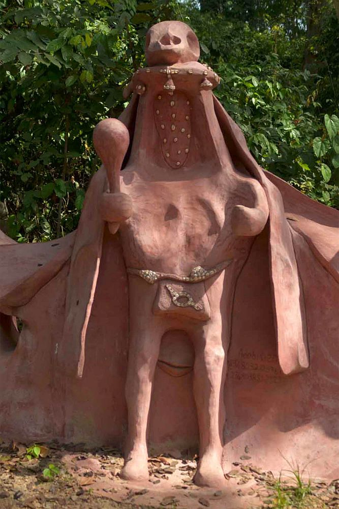 A vaguely human-shaped artwork, appearing to be made out of reddish clay. It has two human legs and wears a bag over its genitals. It has two vaguely-shaped arms coming from an abnormally wide chest. Where the head would be is a short column of sorts, with, on top, what looks vaguely like an animal's skull. It wears a long open cloak and holds something in its hand that looks like a maraca.