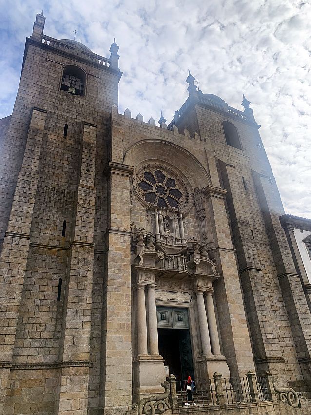 Looking up at the cathedral: built of stone blocks, with two short square towers, each almost as wide as the center entryway. Above an entrance flanked by two pillars on each side is a round window.