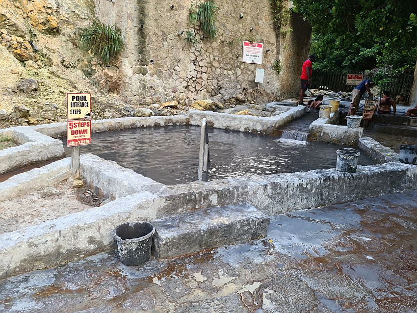 The sulphur bath in the picture is a pool of brown water edged by a simple concrete wall. A channel leads the water into it, visible behind the pool, and also edged with low concrete walls. Signs read "Pool entrance steps" and "5 steps down". Part of the next channel leading the water to the next pool is visible on the left, and plastic buckets stand next to or on the walls here and there.