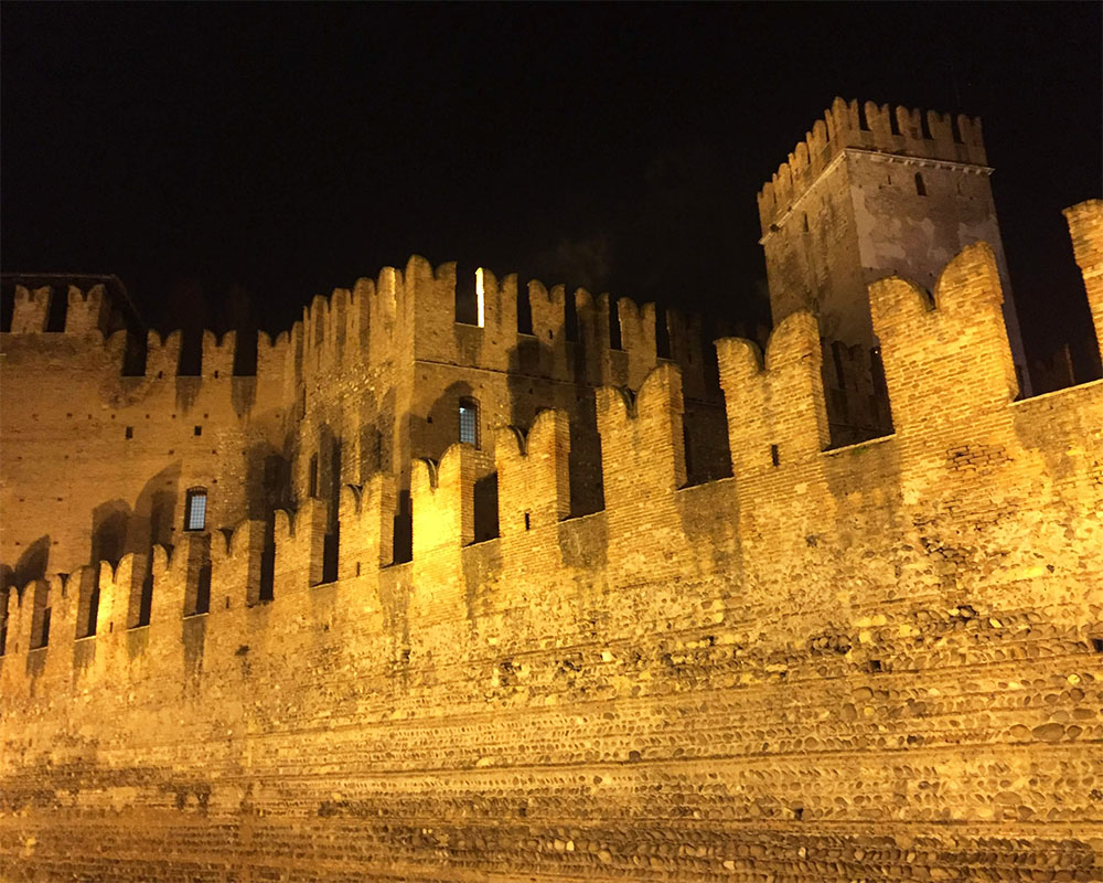 A nighttime view of a length of stone and brick-striped wall, a fortification. It has crenellations, and each upward part of the crenellations has a cleft in its center. Beyond that a building is dimly visible, also stone and brick and also with crenellations along its roof.