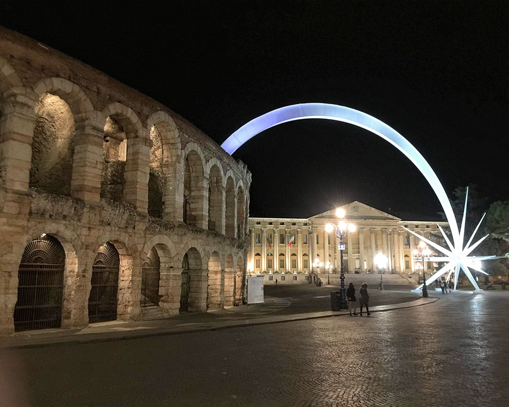 A nighttime shot with, on the left, the rounded wall of the Arena di Verona, a Roman amphitheater with 2 stories, each a row of rounded archways. Beyond that, the Palazzo Barbieri: white with classical columns lining the front. Between the two, a lighted sculpture: a high arch, lit white, with a large white star where it touches the ground.