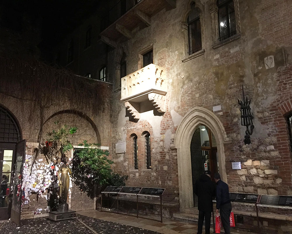 A nighttime shot: walls of stones with some brickwork around the windows. A small balcony is lit up on the upper floor with a small doorway onto the balcony and two small arched windows below it: Juliet's balcony. On the ground below and to the left of the balcony is a full-sized statue in metal, I think, of a woman in flowing dress, presumably Juliet. Behind her are the wall are pinned many small pieces of paper.