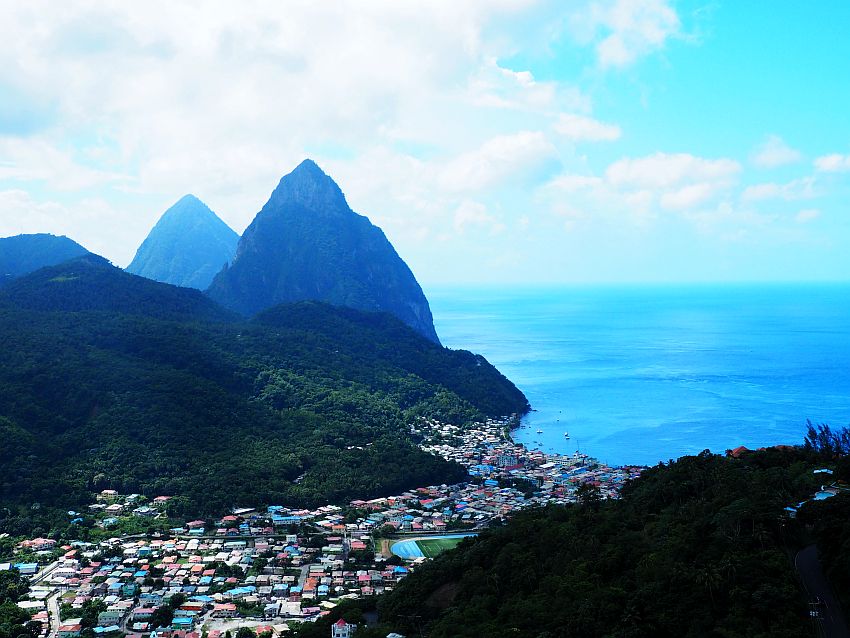 A view from a hill looks across a valley. In the valley a lot of houses, quite close together, in bright colors. They extend up the valley and also down right to the sea on the right. The sea is deep blue. A forest-covered hill rises on the other side of the valley and, beyond that, two taller pointed hills, one partly behind the other.