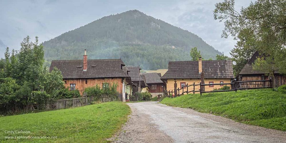 A view of Vlkolínec village from a road entering it: small cottages with shingle roofs on each side of the little road, a tall mountain in the background.