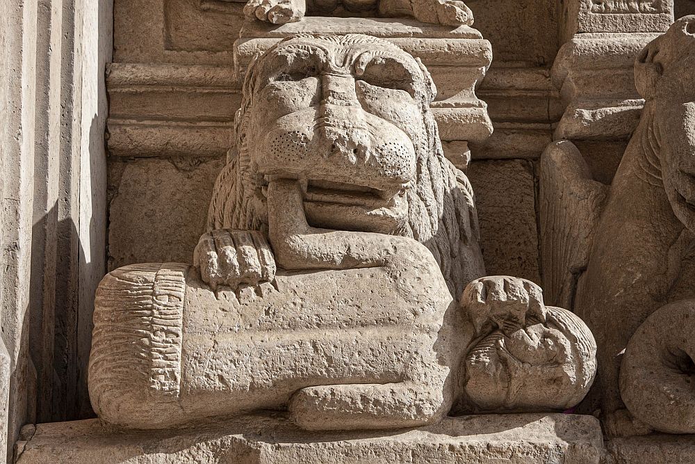 A rather primitive carving: a lion faces the viewer, only its face and paws visible, because they are on top of a person, lying on his side. The man is dead or injured, and the claws of the lion visibly dig into the man's waist and face.