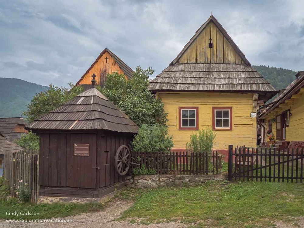 A house in Vlkolínec village: wood shingle roof, yellow wooden walls with dark red trim around the windows. In front is a small outbuilding of dark unpainted wood.
