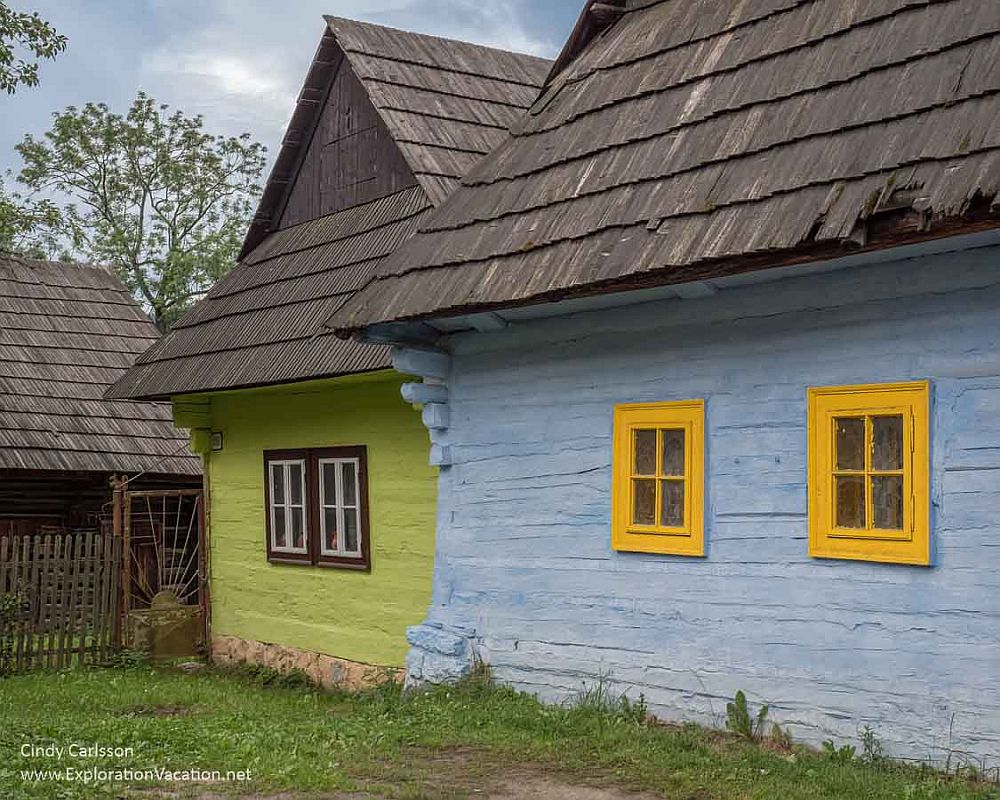 Two of the houses in Vlkolínec UNESCO site, one painted lime green with brown window frames and one painted light blue with bright yellow window frames. Both have wood shingle roofs.
