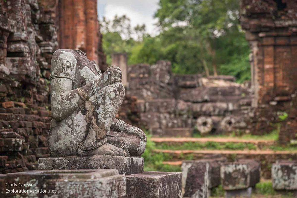 A lichen-covered statue, missing a head, sites with one knee against the pillar it sits on, the other knee up, its hands on the knees. At My Son Sanctuary in Vietnam.