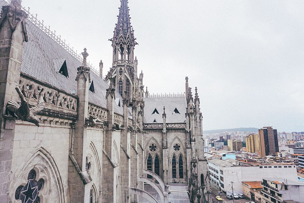 A view along the side of the Basilica in Quito, Ecuador: gothic arches and a very ornate gothic tower, with a bit of the larger city visible in the background.