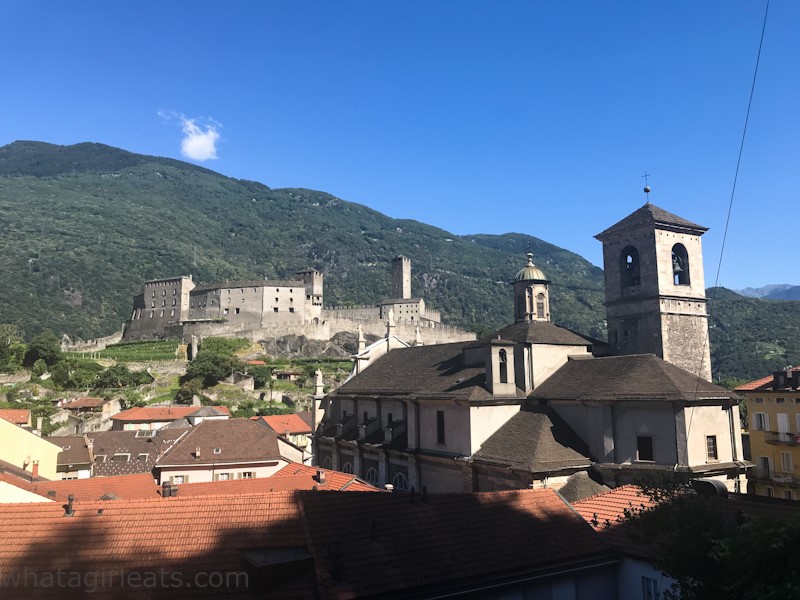 In the foreground, a church with a square bell tower. In the background, on a hill, the castle Castelgrande in Bellinzona. Beyond that, a higher mountain