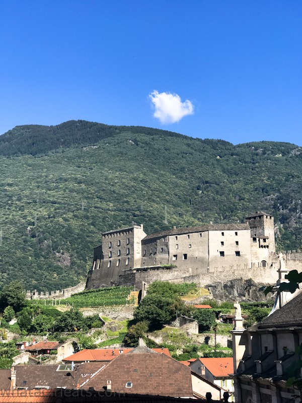 Castelgrande in Bellinzona as seen from below: the castle buildings are simple, with small windows, surrounded by a crenellated wall. Behind the castle, a much higher hill.