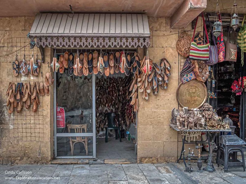 A shop in historic Cairo: One side of the double glass door is open. Leather shoes like mocassins hang on the wall around and above the door. Part of the next shop is visible, and has a table in front covered with small carvings, colorful bags hanging above the table.