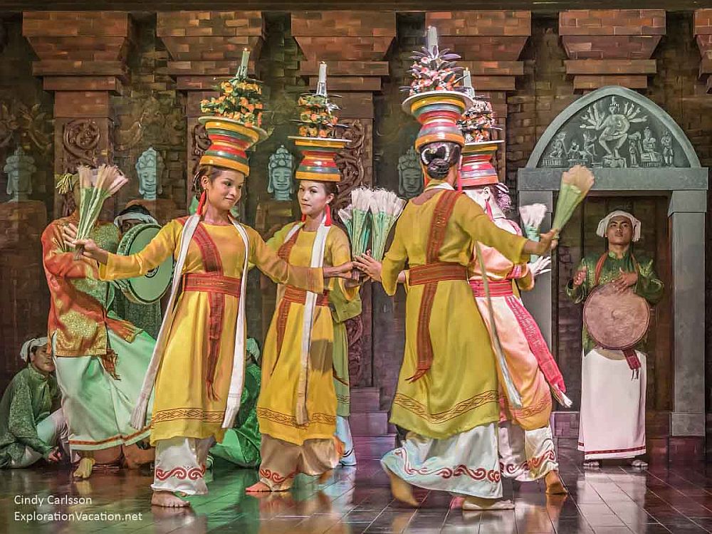 Cham dancers at My Son Sanctuary in Vietnam: women wear yellow dresses with wide white trousers visible underneath, and tall ornate headdresses. They are dancing in a circle, one hand touching in the center.