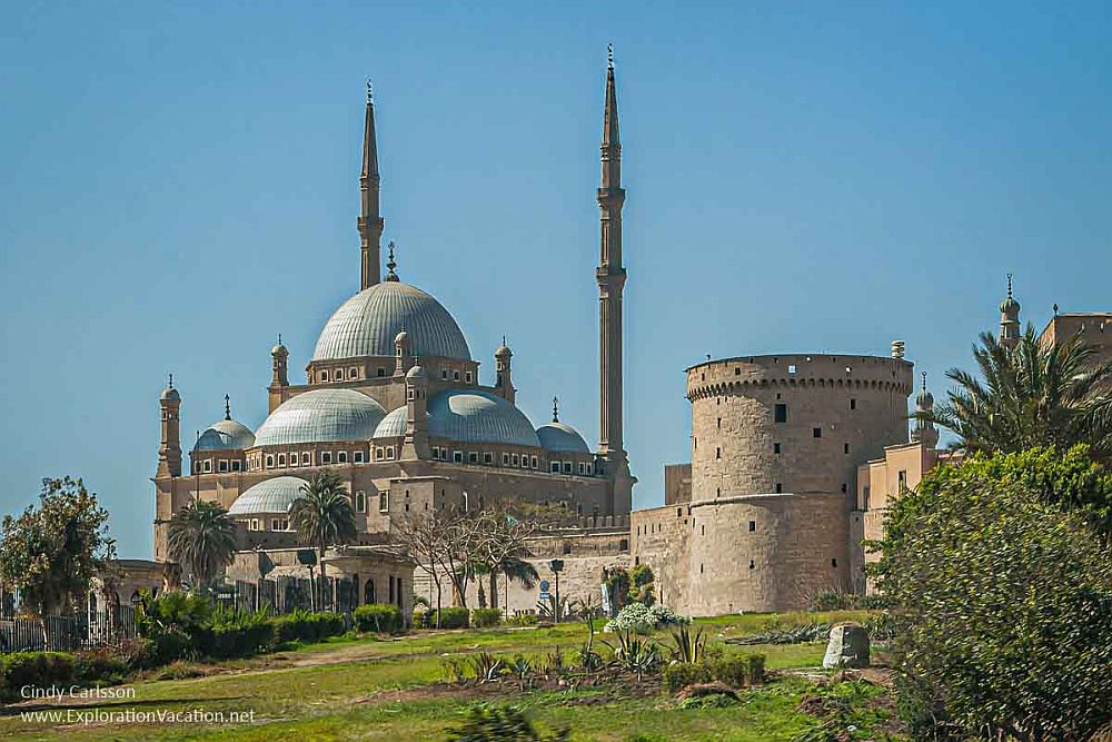 A white mosque with gray domes and very tall slender minarets. Beside it is a structure that looks like a castle's keep, round with crenellations around the top.