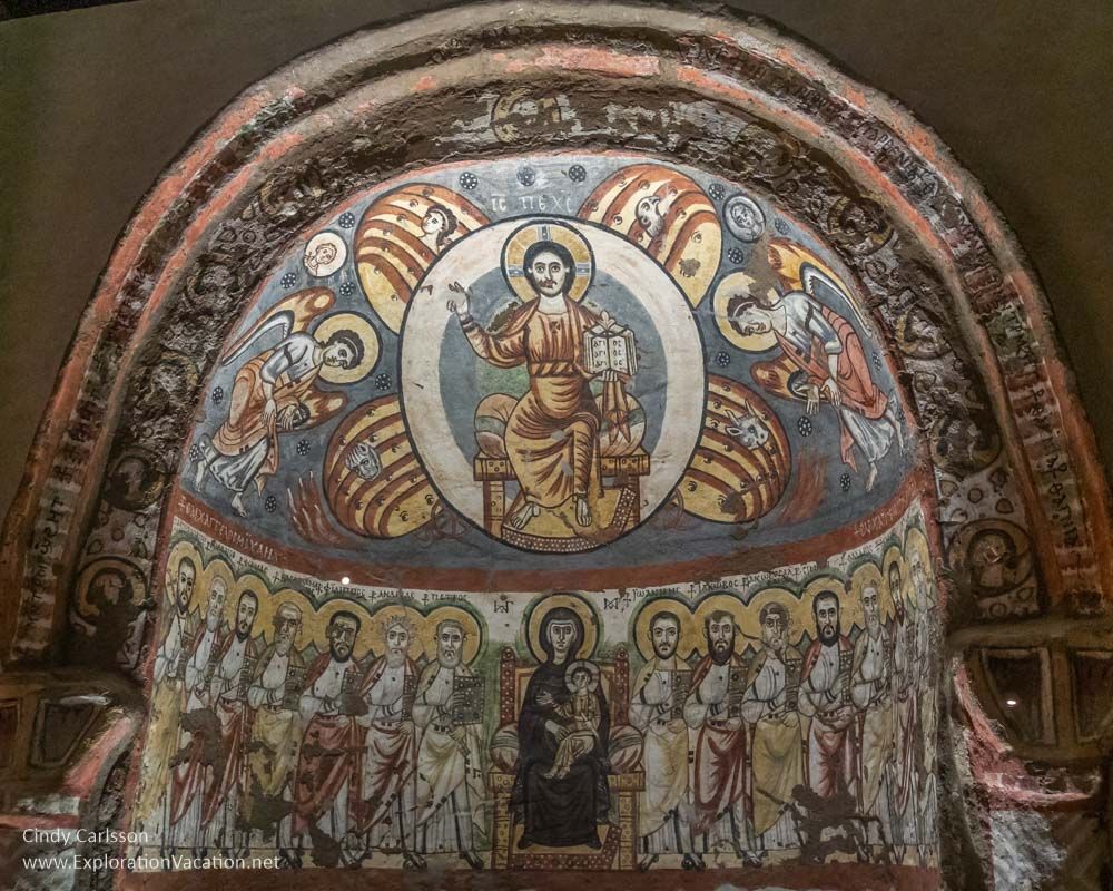 A painted wall with medieval-style Christian images: A saint? or Jesus, with a halo in the center top, and angel on either side of him. Below, the Madonna and Child with the Apostles on either side.