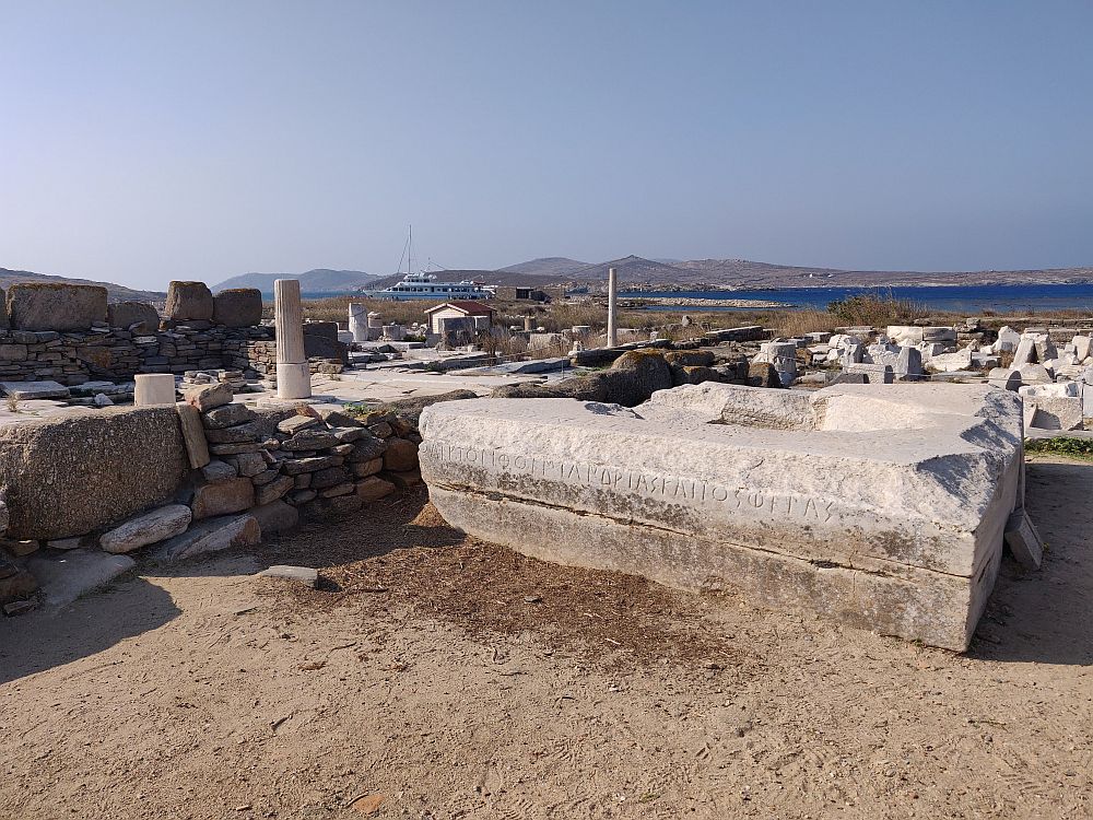 A view over the ruins at Delos: low walls, broken pillars. In the distance, blue water and land beyond that.