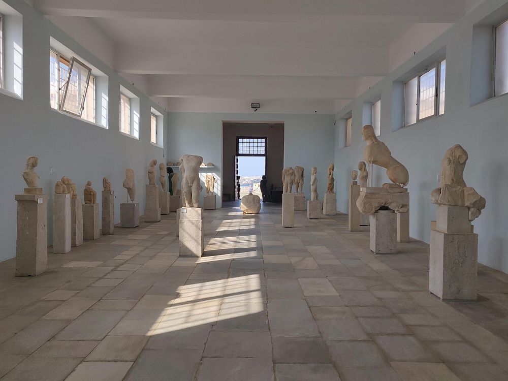 A light-filled hall in a museum. Both side walls are lined with pedestals, and on each pedestal is a statue, most of them eroded and missing parts.