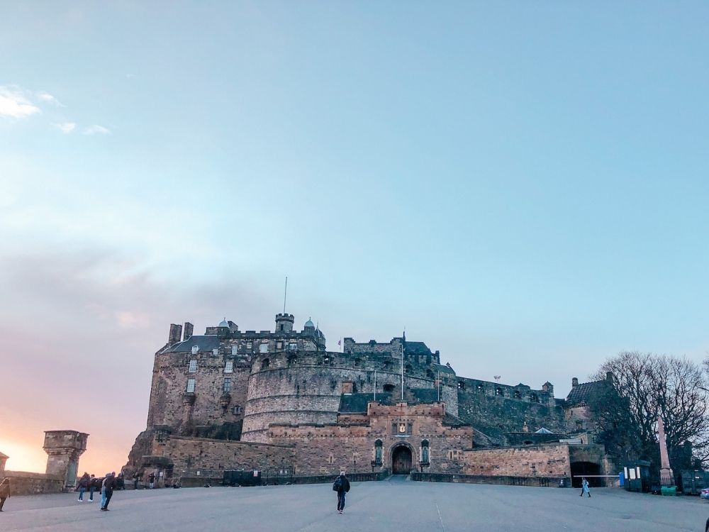 A view of Edinburgh castle, with a sunset sky behind it. The castle has an arched entryway and much higher walls behind the entry, with rounded walls and crenellations.