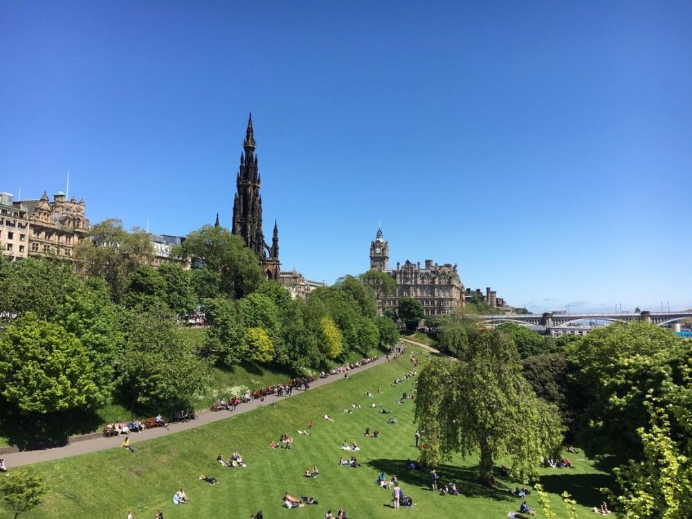 A view of Edinburgh with a grassy slope in front, people sitting on it, a tall church tower and several other buildings in the background.