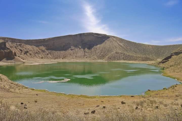 A view of a lake with very dry-looking hills around it. The water is greenish and a flock of flamingos makes a pink spot in it.