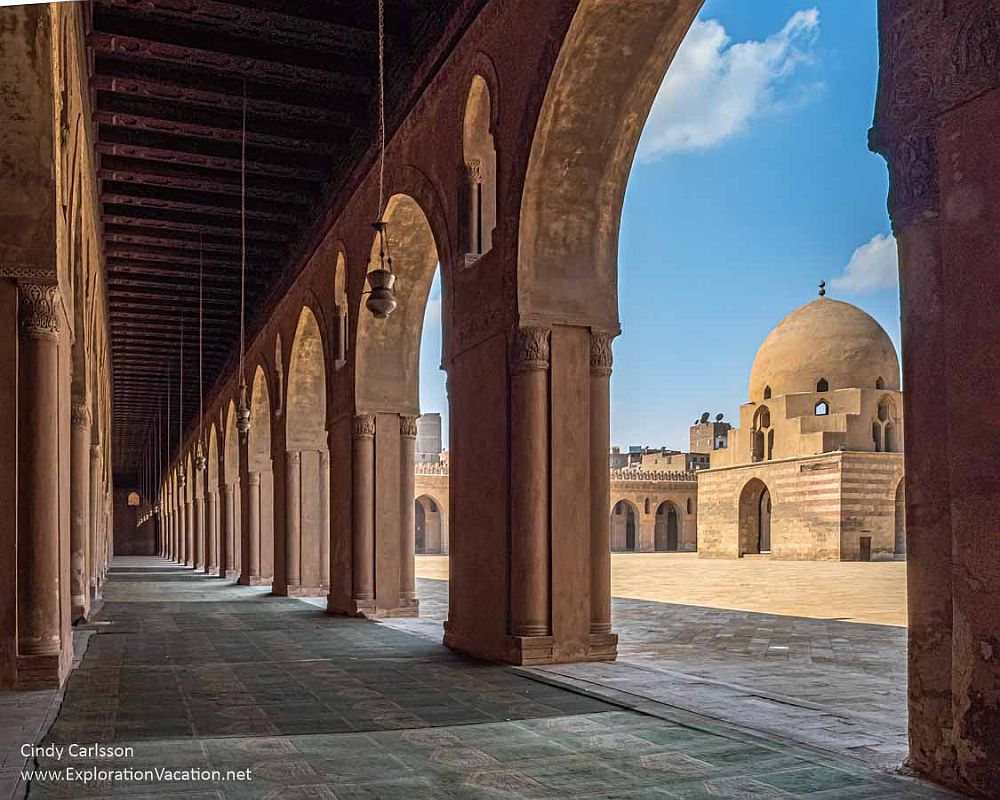 A portico straight ahead, with archways held up by a long line of square pillars on the right. Across a flat open space, a building with more arches and a domed roof.