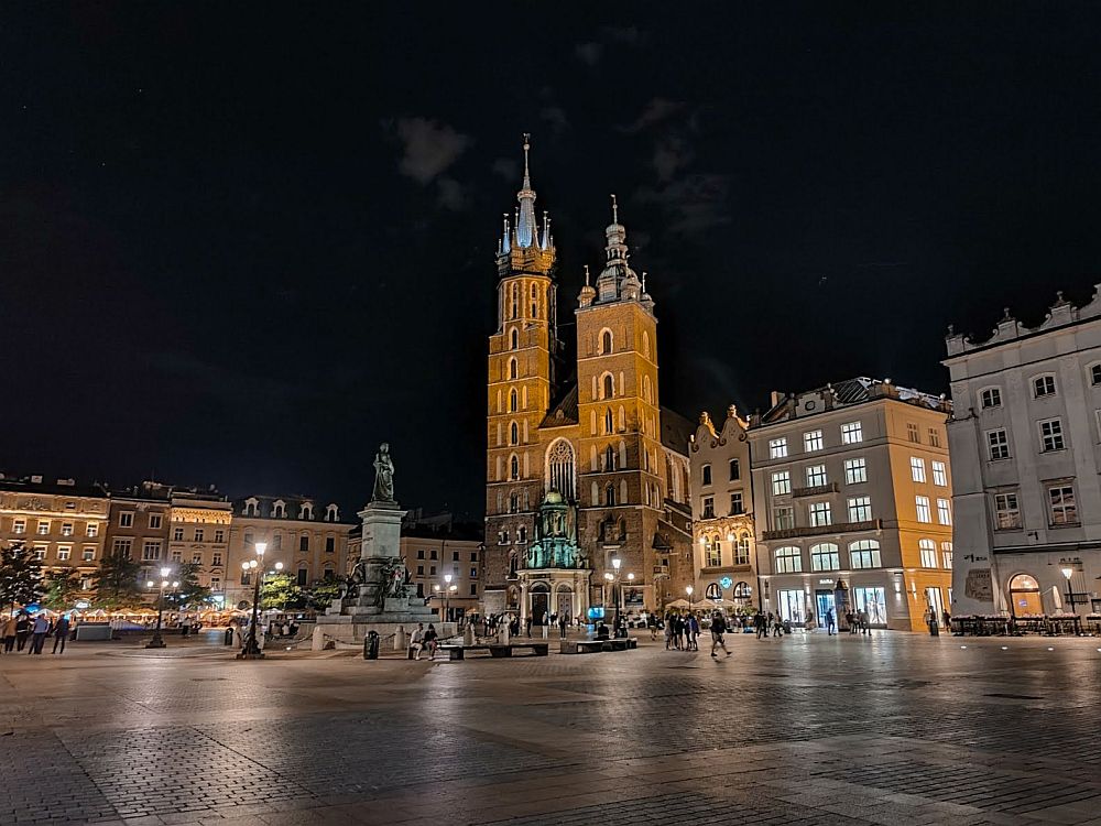 A night seen on a market square in Krakow, Poland: a statue in the square, a church with two uneven spires among several buildings shown on two sides of the square. 